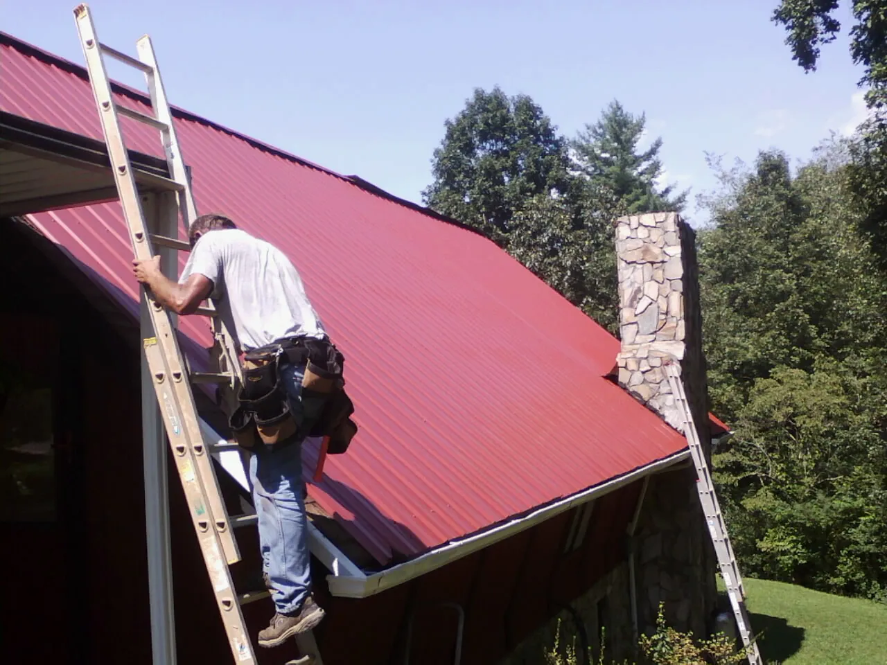 Roofer on a ladder beside a red metal roof and stone chimney — Soffit & Fascia Repair by Roofing Brothers in Fountain Run
