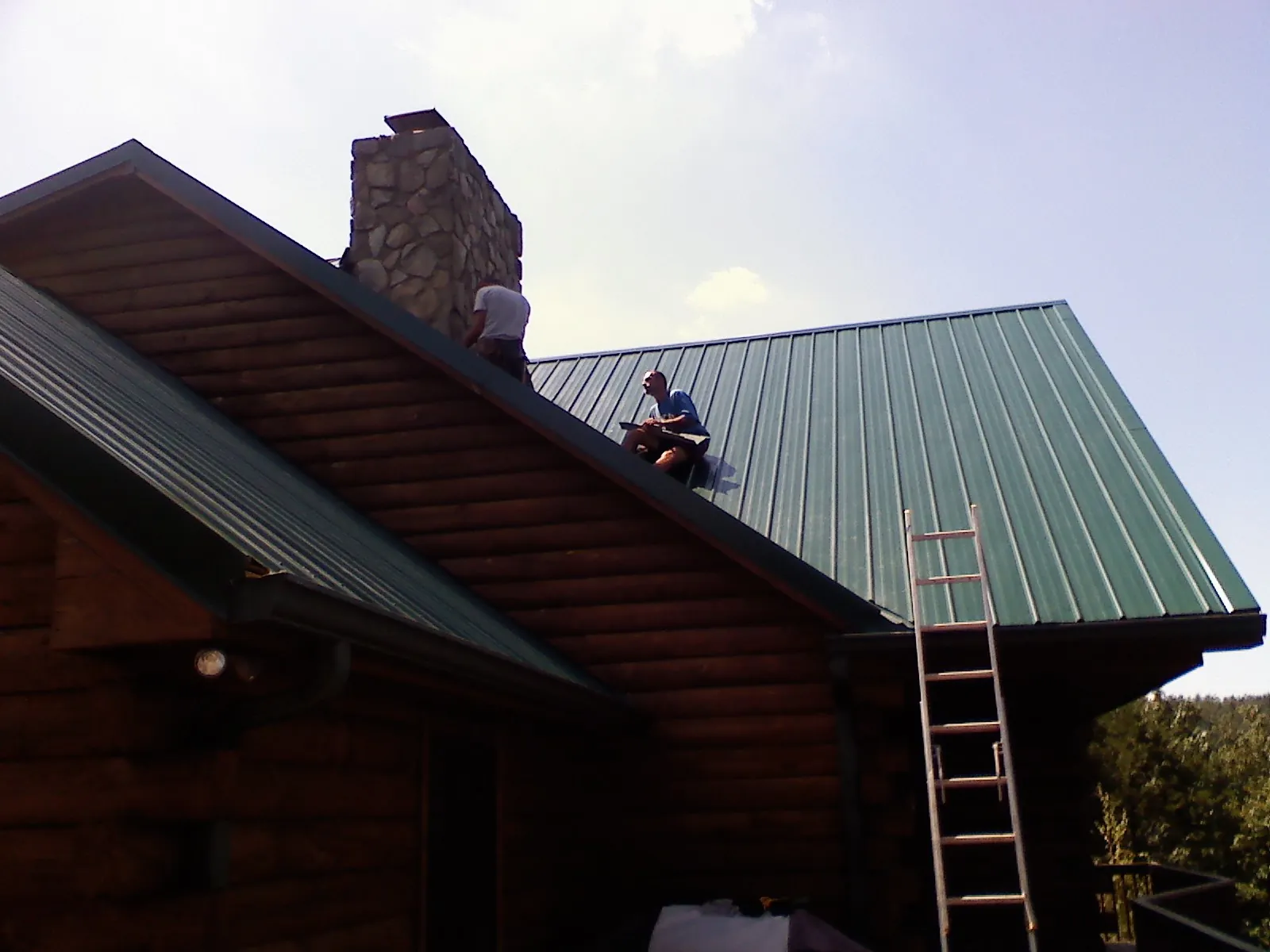 Roofers installing green metal panels beside a stone chimney on a log home in Fountain Run